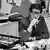 Yves Saint Laurent. Man with glasses in a pin-striped suit sits at a desk littered with fashion related items.