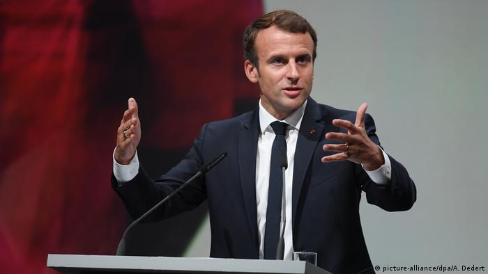 Emmanuel Macron standing at a podium, publicly speaking, gesturing with his hands.