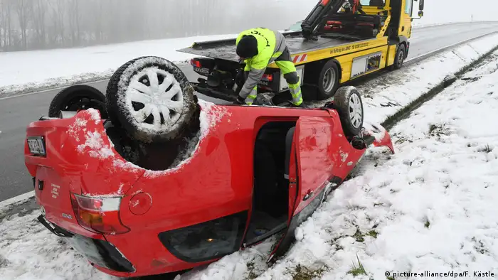A car turned upside-down on the side of the street, a tow-truck in the background, and snow on the ground.
