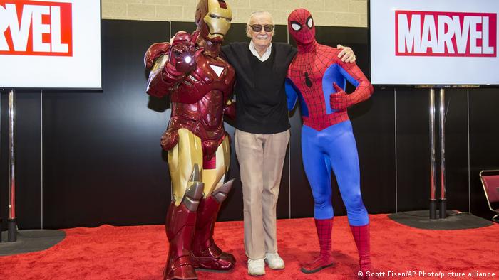 Ein älterer Mann steht auf dem roten Teppich zwischen den Comicfiguren Spiderman und Iron Man.