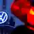 Logomarca luminosa da Volkswagen. Ao lado, em primeiro plano, desfocado, o sinal vermelho de um semáforo. 