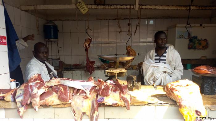 Two men sitting at a market booth, with large pieces of meat on a counter in front of them.