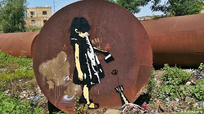Auf einer großen rostenden Metallscheibe ist ein Mädchen abgebildet, das aus einem kleinen Gefäß Wasser auf eine Blume gießt