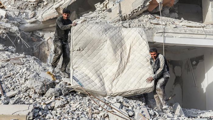 Two men carrying a mattress through the rubble
