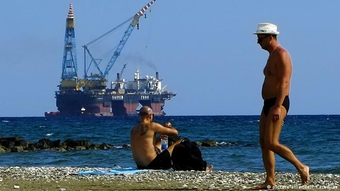 A man walks as an other man sits on a beach as a drilling platform is seen in the background outside from Larnaca port