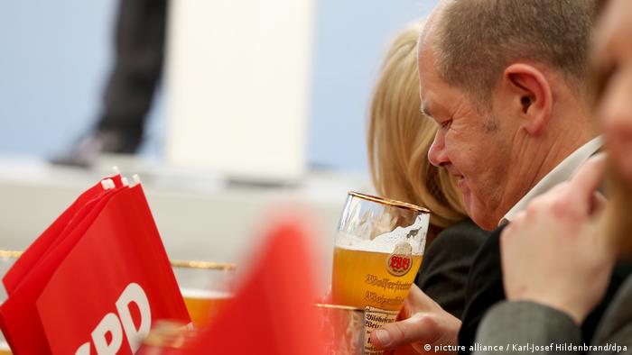 Olaf Scholz verzieht das Gesicht in sein Bierglas blickend, daneben SPD-Fähnchen (Foto: Picture Alliance)