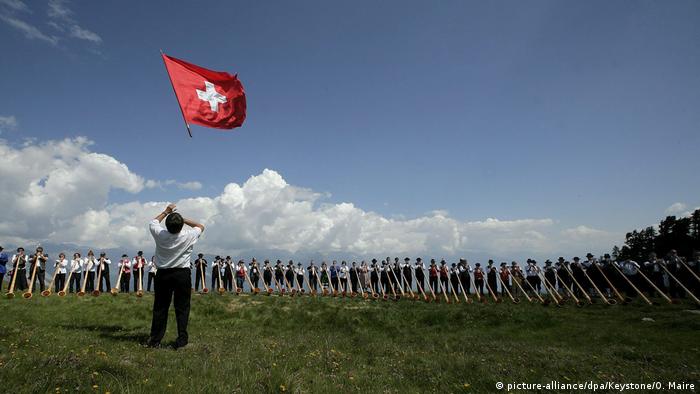 Schweizer Fahne vor einem Panorama aus Alphorn-Bläsern anlässlich des 5. internationalen Alphorn-Festivals in Nendaz in der Südschweiz.