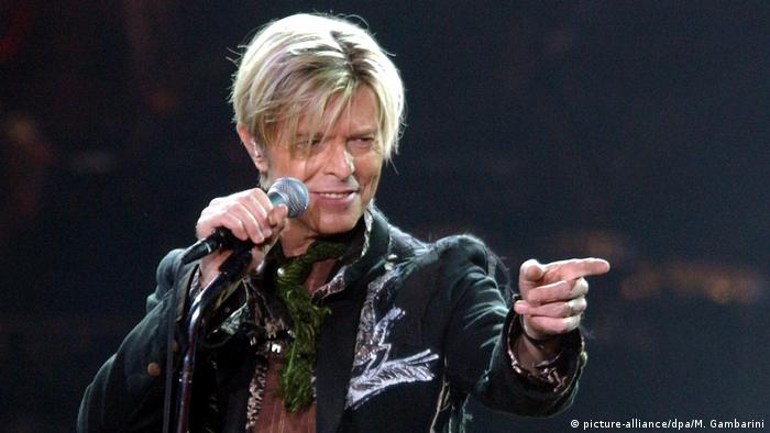 David Bowie in 2003, smiles and points towards the audience while holding a microphone.