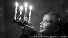 Schwarz-Weiß-Foto: Ein Mädchen hält einen kleinen Holzadventkranz mit leuchtenden Augen hoch, auf dem vier dünne Kerzen brennen