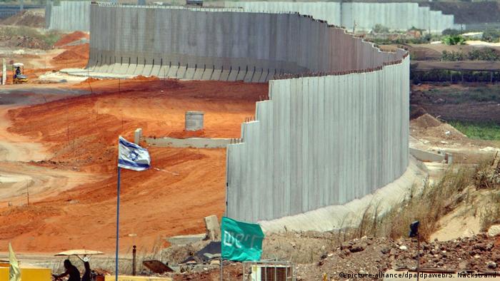 A concrete wall under construction winds its way along a base in the desert while an Israeli flag waves in front (picture-alliance/dpa/dpaweb/S. Nackstrand)