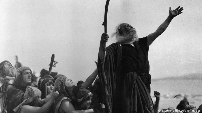 Scene from the 1923 film The Ten Commandments featuring an actor playing Moses reaching toward the sky with people behind him.