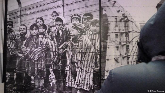 Eine Person, von der nur Kapuze und Schulter zu sehen sind, steht vor einer Ausstellungswand mit einem großen Foto: Darauf sind Kinder in gestreifter, viel zu großer Kleidung hinter einem Stacheldrahtzaun zu sehen