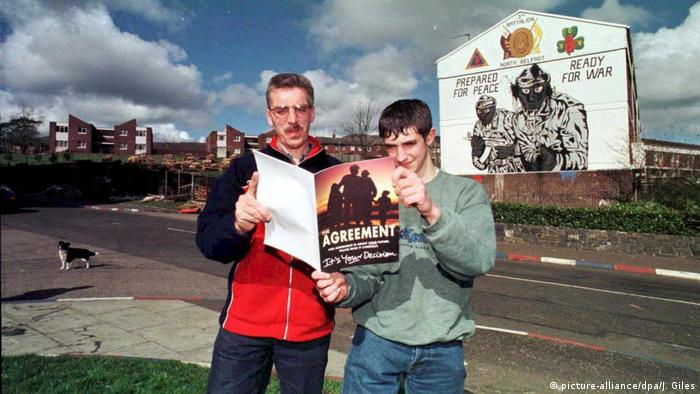 Two men read a copy of the peace agreement in front of a mural that reads 'Prepared for peace, ready for war'
