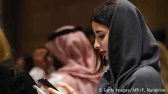 A woman wearing a headscarf is looking at her mobile phone, in the background is a man with a traditional Saudi Arabian headscarf.