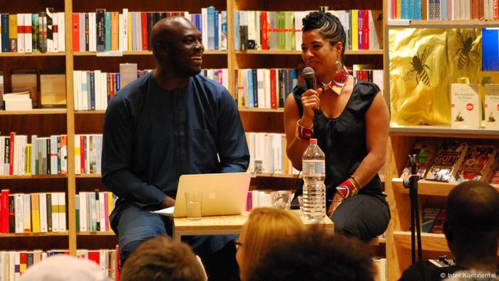 A man and a woman seated at a small table in front of shelves of books, Elnathan John and Olumide Popoola