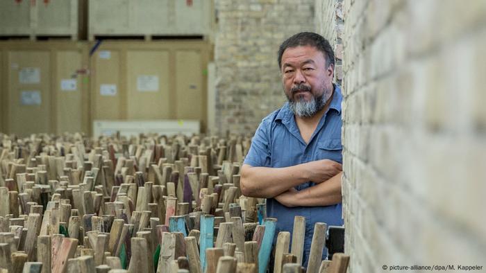 Mit verschränkten Armen steht Aiweiwei in einem blauen Hemd in seinem Studio, umgeben von niedrigen Holzstämmen