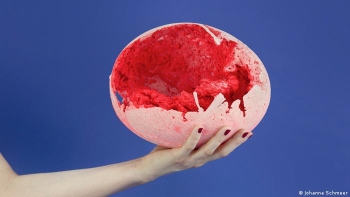 Eine Hand hält ein großes rosafarbenes Ei, in dessen zerbrochener Schale eine rote Masse zu sehen ist. Aus: Bioplastic Fantastic (Johanna Schmeer).