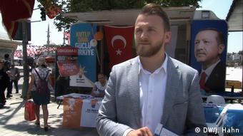 Emin Sarıoğlu stands and looks off camera as Erdogan's face hangs on a poster in the background