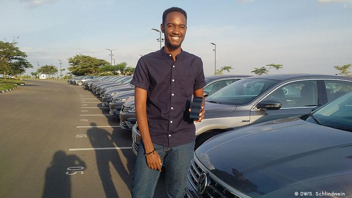 Mpfizi stands in front of a row of cars, holding up his smartphone