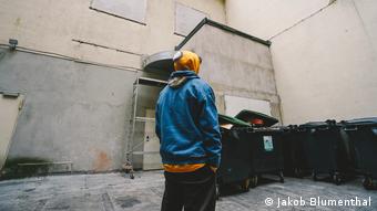 A person wearing a sweatshirt with a kippa on the hood stands near dumpsters