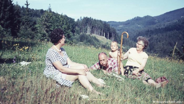Eine Familie sitzt bei Sonnenschein auf einer Wiese in einer bergigen Landschaft.