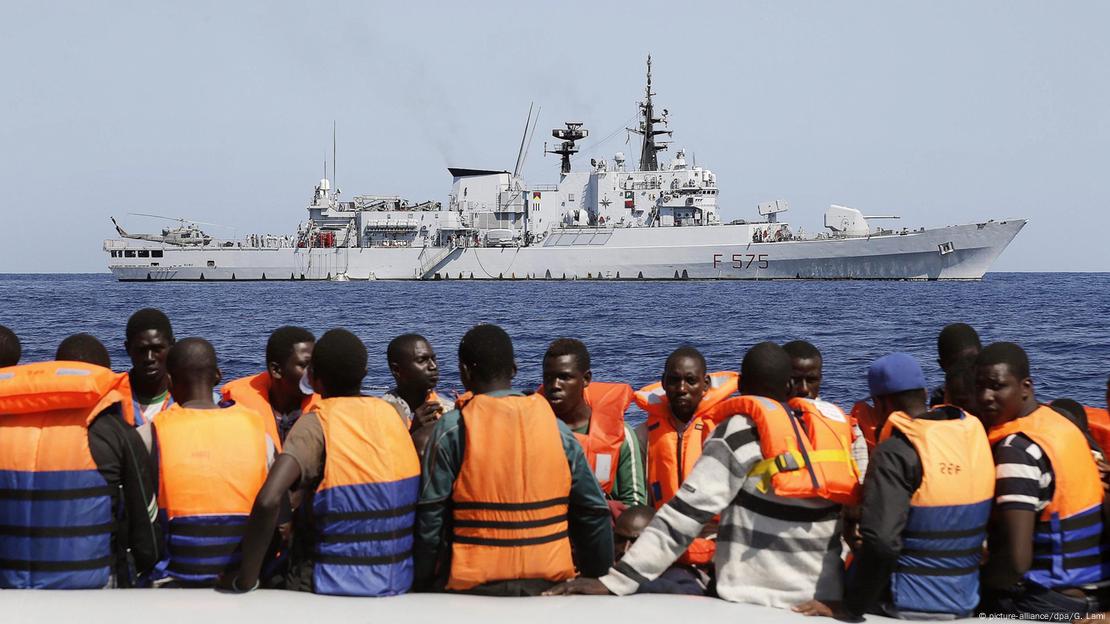 Ein italienisches Marineschiff im Hintergrund, im Vordergrund Männer mit Schwimmwesten (auf einem Boot?)