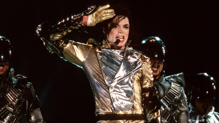 Michael Jackson wearing a gold and silver outfit sings into a microphone. 