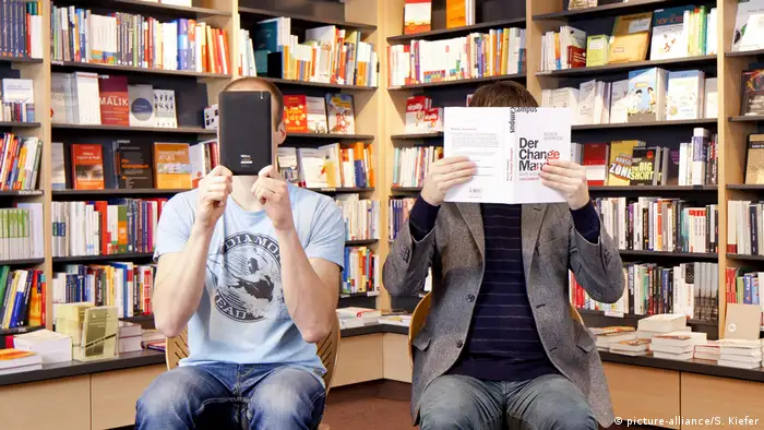 Symbolbild: Ein Mann hält sich ein elektronisches Buch, E-Book-Reader, ein anderer Mann ein konventionelles Buch vor das Gesicht