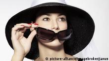 Junge Frau mit schwarzem Hut und schwarzem Kleid hält Sonnenbrille vor das Gesicht | Verwendung weltweit, Keine Weitergabe an Wiederverkäufer.