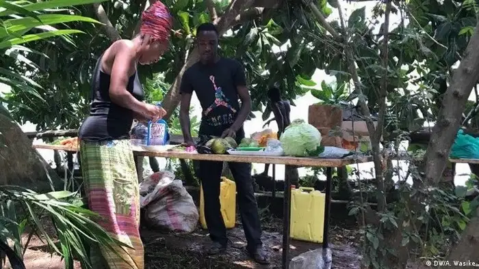 A man and a woman in a pink headwrap stand over a table laden with vegetables under a few trees