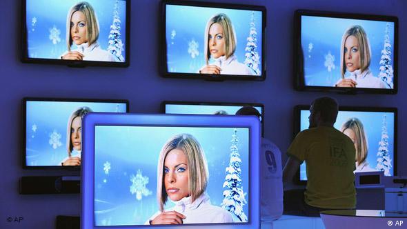 Bir fuar alanında farklı boyutlardaki çok sayıda televizyon ekranı aynı görüntüyü eş zamanlı olarak gösteriyor.