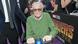 Stan Lee sitting in a cart, with people surrounding him. Stan Lee sitting in a cart, with people surrounding him.