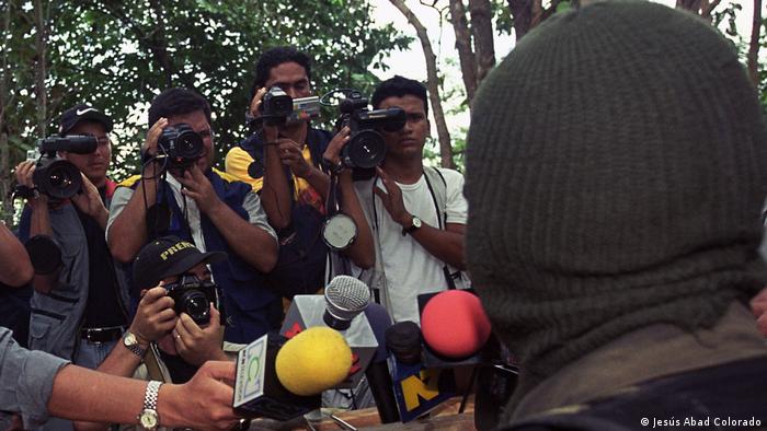 A masked person in front of journalists holding microphones