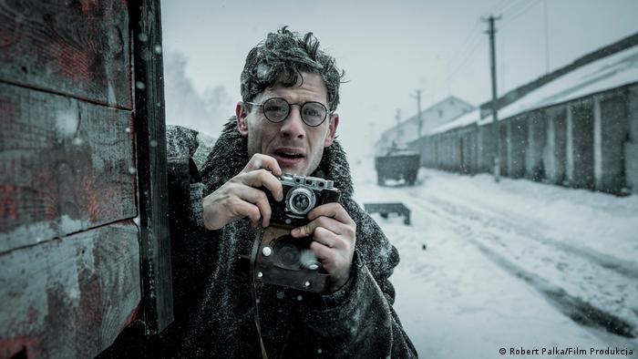 Ein Filmausschnitt aus dem Film Mr Jones zeigt einen Mann mit Kamera in einer verschneiten Landschaft neben einem Zug 
