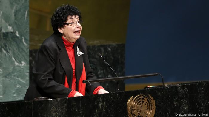 Eine Frau mit Brille in einem dunklem Jackett und roten Pulli steht vor einem Rednerpult und spricht