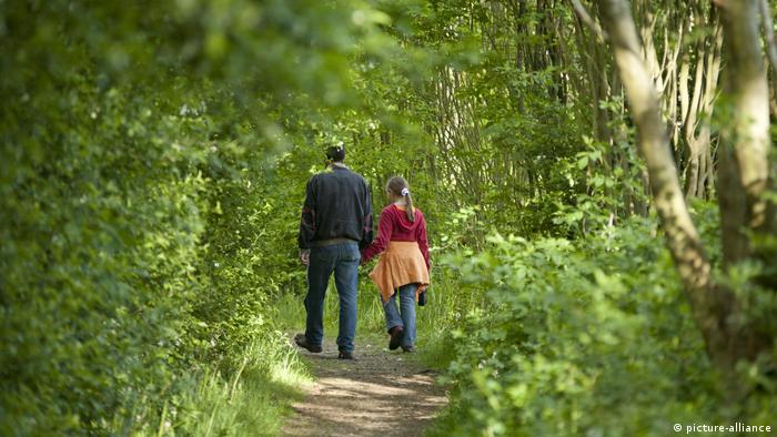 Wir blicken auf ihre Rücken: Ein Mann geht mit einem Mädchen auf einem schmalen Weg durch einen hellgrünen Laubwald. Sonnenlicht fällt durch das Blätterdach (picture-alliance)
