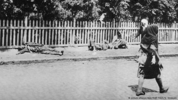 Schwaz-weiß-Fotografie von 1932/1933: Auf dem Gehweg vor einem Gartenzain liegen verhungerte Leichen, zwei Frauen gehen achtlos vorbei