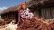 A woman stands in front of a long wooden hut next to a pile of dried plants A woman stands in front of a long wooden hut next to a pile of dried plants