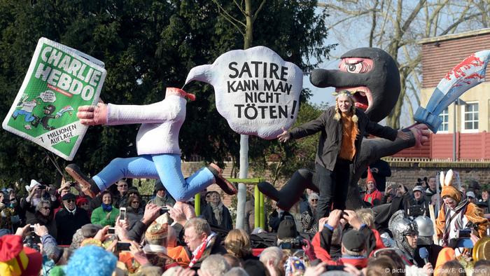 Eine geköpfte Figur auf Tillys Wagen hält eine Charlie Hebdo-Ausgabe in der Hand und sagt laut Sprechblase Satire kann man nicht töten!, dabei verfolgt sie eine maskierte Figur mit blutverschmiertem Schwert
