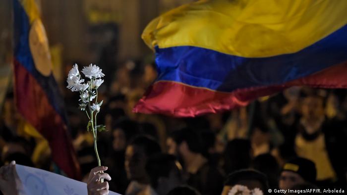 Foto simbólica de una persona con una flor blanca en la mano y la bandera de Colombia en el fondo.