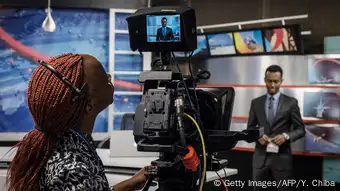 in a tv studio, a woman looks at the picture of a news anchor on a camara display 