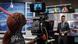 in a tv studio, a woman looks at the picture of a news anchor on a camara display 