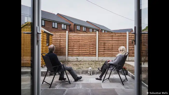 A man and a woman sitt in garden chairs on a patio in a yard walled off by a fence with tenement hosues in the background (Copyright: Sebastian Wells)