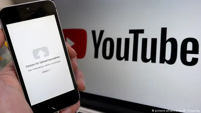 Mobile phone showing upload symbol with YouTube logo on computer screen behind