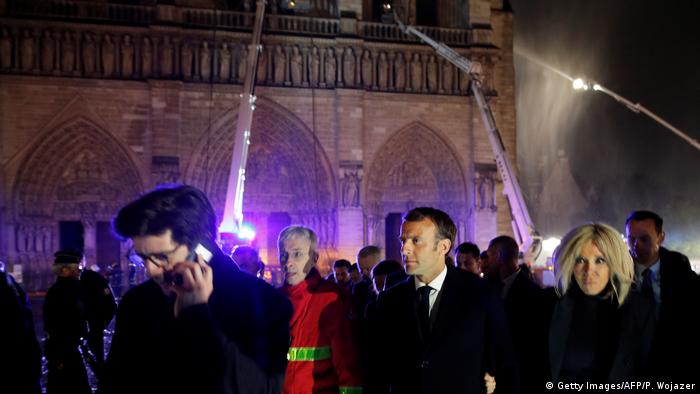 French President Emmanuel Macron and his wife Brigitte visiting the scene in the evening, with firefighters in the background. 