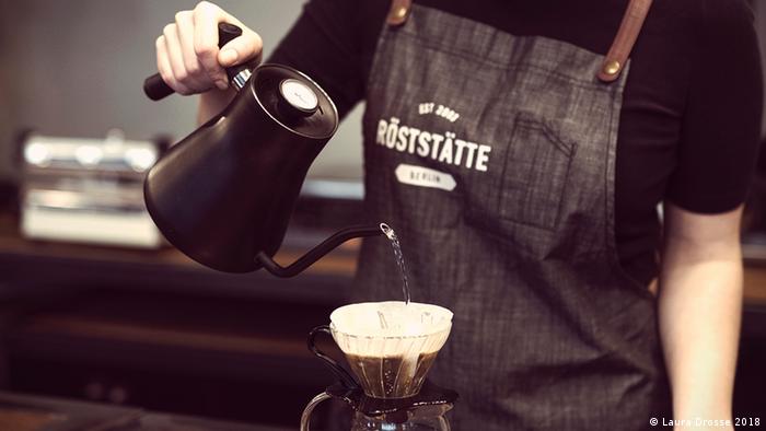 Hand gießt Wasser aus einer schwarzen Kanne in einen Kaffeefilter ein (Foto: Laura Drosse 2018).