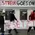 Three students carry a banner under a sign that says "The Streik Goes On"