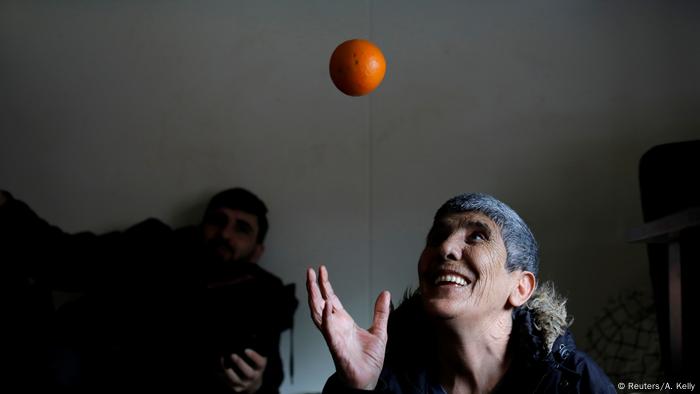 Der Iraner Karim Azizi wirft eine Orange in die Luft und schaut ihr mit einem großen Lächeln nach. Hinter ihm sitzt ein weiterer Bewohner des Abschiebelagers Kærshovedgård und schaut ihm zu.