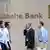 Quatro homens e uma mulher caminham diante de parede com logo do Deutsche Bank