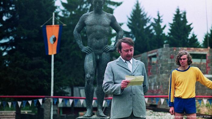 Filmstill aus Der nackte Mann auf dem Sportplatz mit Sportler und weiterem mann auf einem Fußballplatz mit Fahne und großer Steinfigur